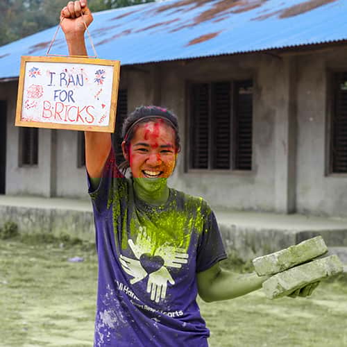 Volunteer-Smiling-Fundraise-Holi-Run.jpg A smiling volunteer, covered in paint, holding sign that shows her fundraising efforts in the Nepal Holi Run