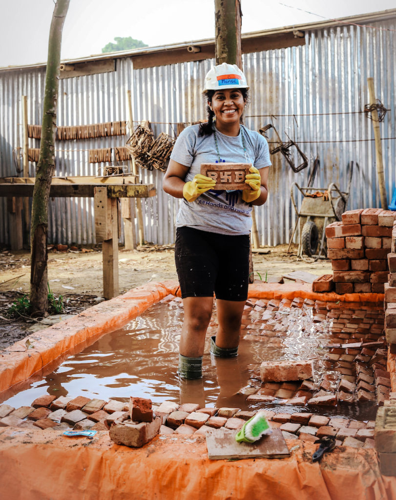 Female Nepal volunteer stands smiling stacking bricks. 